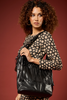 [COLOR: BLACK LEATHER]A woman with curly brown hair models a large black faux-leather shoulder bag with fringe detailing, set against a solid dark red studio background.