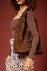 [color: chocolate] A woman wearing a brown long-sleeve top, carrying a large brown suede shoulder bag with braided trim and a dark metal buckle, against a solid dark red background.
