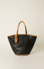 A black leather tote bag with braided tan handles and a matching tassel charm, photographed against a neutral light beige background.