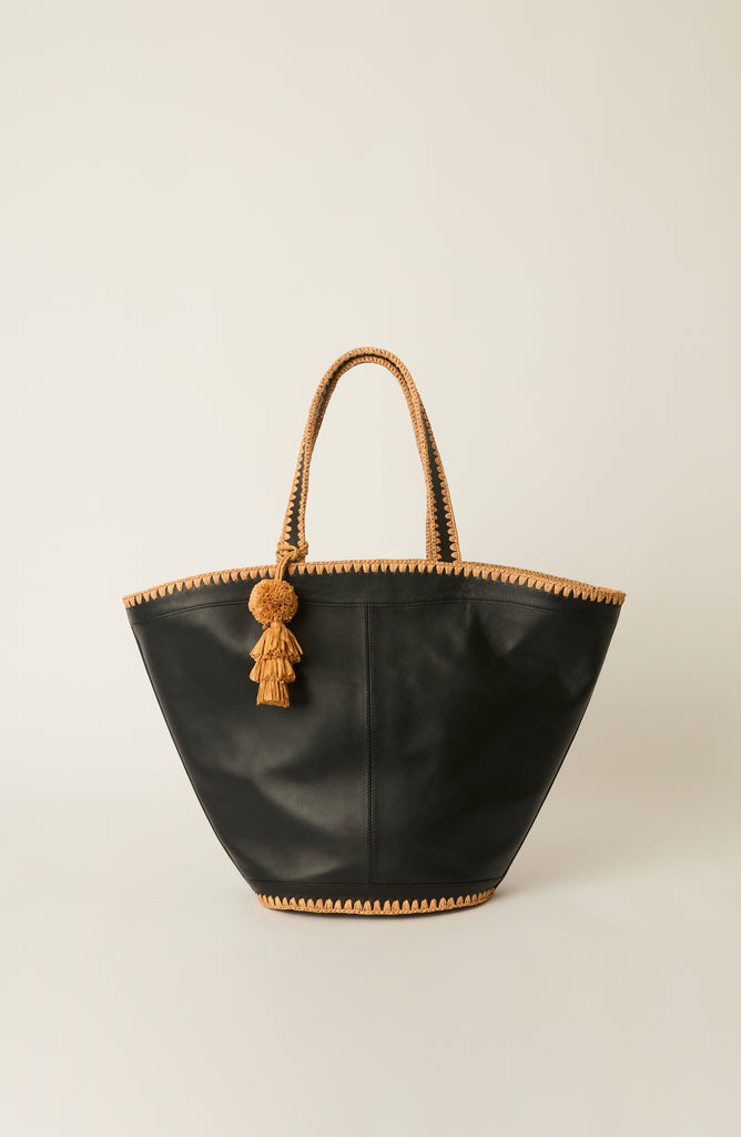 A black leather tote bag with braided tan handles and a matching tassel charm, photographed against a neutral light beige background.
