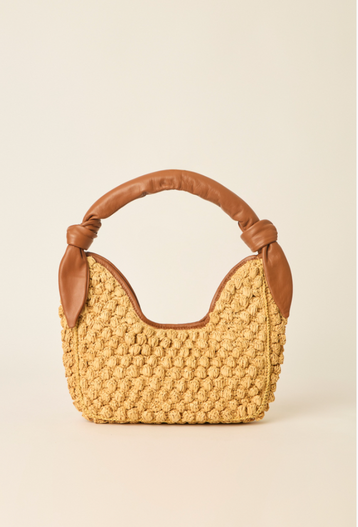 A small woven straw handbag with a tan leather handle and trim, photographed against a plain warm off-white background.