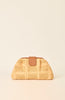 A small woven straw clutch bag in a warm beige color with a smooth tan leather strap closure, set against a solid cream background.