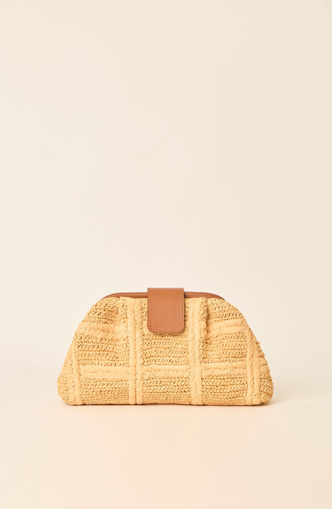 A small woven straw clutch bag in a warm beige color with a smooth tan leather strap closure, set against a solid cream background.