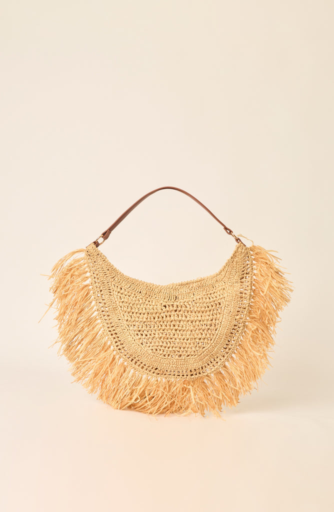 A natural straw crescent-shaped shoulder bag with long fringe detailing and a brown leather handle, set against a solid cream background.