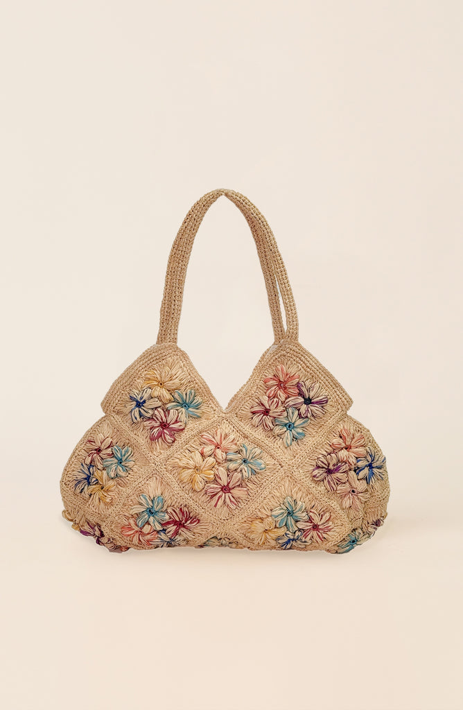 A beige woven straw handbag with colorful embroidered floral patterns and two top handles, set against a solid cream background.