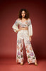 A model with curly hair stands against a solid dark red studio background wearing a matching two-piece set consisting of a long-sleeve crop top and wide-leg pants, both in an off-white base with a colorful tropical leaf print.