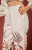 A close-up of a model wearing high-waisted off-white trousers with colorful embroidered tropical palm leaves in shades of blue, orange, and green. The model's hand is resting on the hip pocket, and the background is a solid dark red.