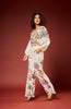 A woman modeling a matching two-piece outfit consisting of a long-sleeve crop top and wide-leg pants, both in white with a colorful tropical leaf print, set against a solid dark red studio background.