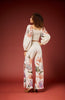 A woman stands with her back to the camera wearing a matching two-piece outfit consisting of a smocked crop top and wide-leg trousers, both in an off-white base with a colorful botanical leaf print. She is posed against a solid dark red studio backdrop.
