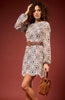 Woman wearing a patterned dress with a brown belt and holding a brown handbag against a red background
