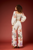A model is shown from the back wearing a matching two-piece outfit consisting of a white textured crop top with dramatic puff sleeves and wide-leg pants featuring a colorful botanical leaf print, set against a solid dark red studio backdrop.