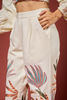 A close-up of a model wearing off-white wide-leg trousers with colorful embroidered tropical palm leaves in shades of teal, orange, and green. The model is also wearing a cream-colored cropped top, set against a solid dark reddish-brown background.