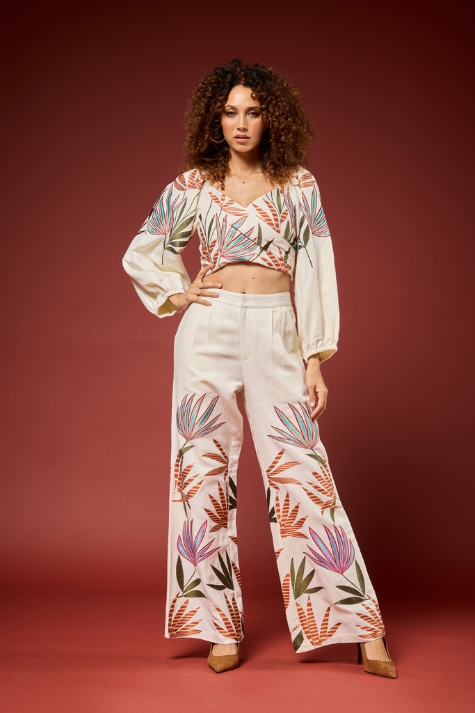 A model stands against a solid dark red studio background wearing a matching two-piece set consisting of a long-sleeve wrap-style crop top and high-waisted wide-leg trousers, both featuring a colorful botanical palm leaf print.