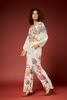 A model wearing a matching two-piece lounge set with a tropical palm leaf print, featuring a long-sleeve crop top and wide-leg pants, posed against a solid dark red studio backdrop.