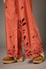 Close-up of a person wearing wide-leg terracotta orange pants with leaf-shaped cutout embroidery along the hems, paired with tan espadrille wedge sandals against a neutral gray studio background.