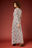 A woman modeling a long-sleeved maxi dress with a detailed geometric and floral pattern in blue, brown, and cream, shown from a side-back angle against a solid dark red studio backdrop.