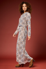 A woman modeling a long-sleeve maxi dress with a detailed geometric medallion pattern in blue, orange, and brown on a cream base, paired with brown suede boots, set against a solid dark red studio backdrop.