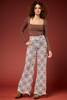 A woman modeling a brown long-sleeve scoop-neck top and high-waisted, wide-leg pants with a colorful geometric medallion pattern in blue, orange, and cream, set against a solid dark red studio background.