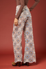A model is shown from the back wearing high-waisted, wide-leg pants with an intricate geometric and floral pattern in beige, brown, blue, and white. The background is a solid terracotta color.