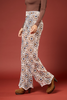 A model wearing high-waisted, wide-leg pants with a detailed geometric and floral mandala pattern in beige, brown, blue, and white. The pants feature a scalloped lace hem and are styled with a brown long-sleeve top and brown suede boots against a solid rust-red studio background.