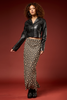 A model stands against a dark red studio backdrop wearing a black cropped leather-look button-up shirt and a long black skirt with a beige circular pattern, paired with black boots.