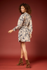 A woman modeling a beige long-sleeve mini dress with a navy blue abstract floral pattern, paired with brown suede ankle boots, set against a solid dark red studio backdrop.