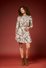 A woman with curly brown hair models a long-sleeve, knee-length cream dress with a navy blue abstract floral pattern, paired with brown suede ankle boots, against a solid dark red studio backdrop.