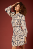 A woman with curly brown hair models a long-sleeve mini dress featuring a beige and navy blue abstract floral pattern, posed against a solid dark reddish-brown studio background.