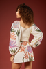 A model with curly brown hair is shown from the back wearing a cream-colored mini dress with a smocked bodice and voluminous sleeves, decorated with a colorful tropical leaf pattern. The background is a solid dark reddish-brown studio backdrop.