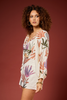 A woman with curly brown hair models a long-sleeve, short romper-style dress with a tropical floral print. The garment has a cream base with orange, green, and purple botanical motifs, set against a solid dark reddish-brown studio background.