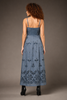 A woman viewed from the back wearing a long blue denim maxi dress with thin straps, a fitted bodice, and ornate floral embroidery concentrated along the lower skirt, paired with black high heels against a plain taupe studio background.