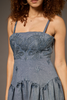 A close-up of a model wearing a blue denim-style dress with floral lace applique on the bodice and thin shoulder straps, shown against a plain gray studio background.