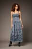 A woman modeling a sleeveless, floor-length blue dress with black floral embroidery and black ankle boots, photographed against a plain taupe studio background.