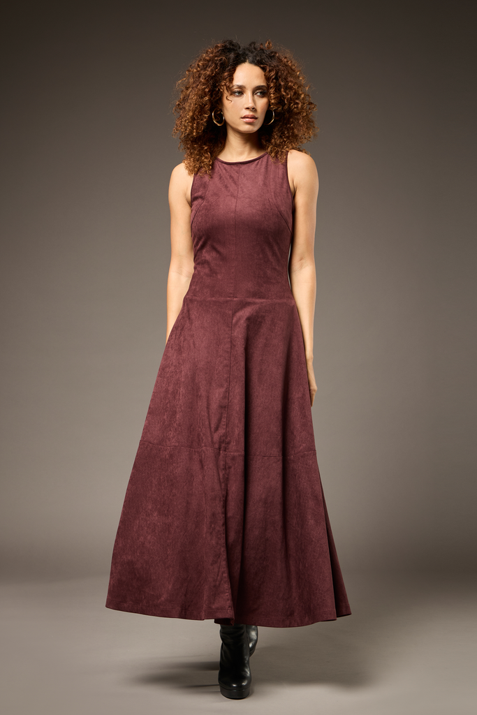 A woman modeling a sleeveless, high-neck, floor-length A-line dress in a deep burgundy or maroon suede-like fabric, paired with black ankle boots, against a dark gray studio background.