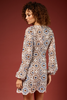 A woman with curly brown hair is shown from the back wearing a long-sleeve bohemian-style mini dress with an intricate geometric mandala pattern in beige, brown, blue, and white, set against a solid dark reddish-brown studio background.