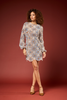 A woman modeling a long-sleeve mini dress with a detailed geometric mosaic pattern in blue, beige, and brown, set against a solid dark red studio background.