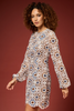 A woman modeling a long-sleeve mini dress with a detailed geometric and floral crochet-like pattern in beige, brown, blue, and white, set against a solid dark reddish-brown studio background.
