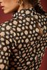 Close-up of a model wearing a black long-sleeve blouse with a beige floral pattern and a high mock neck, set against a solid dark red background.