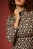 A close-up of a woman wearing a black long-sleeve blouse with a beige and pink floral pattern, paired with large gold hoop earrings, set against a solid dark red background.