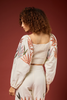 A model with curly brown hair is shown from the back, wearing a cream-colored two-piece set featuring a smocked crop top and matching pants, with long sleeves printed in a botanical leaf pattern of orange, green, and blue. She is posed against a solid dark reddish-brown studio background.