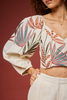 A close-up of a model wearing a cream-colored crop top with voluminous puff sleeves and a vibrant embroidered tropical leaf pattern in orange, teal, and olive green, paired with white trousers against a solid dark red background.