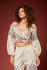 A woman with curly brown hair models a matching two-piece set featuring a cream-colored long-sleeve crop top and high-waisted pants, both decorated with a colorful palm leaf pattern in shades of orange, green, and blue, set against a solid dark red background.