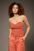 A woman with curly brown hair models a terracotta-colored strapless lace top and matching wide-leg pants against a solid dark gray studio background.