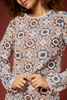 A close-up of a woman wearing a long-sleeve knit top with an intricate geometric mandala pattern in brown, beige, white, and blue, set against a solid dark reddish-brown background.