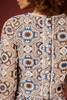 Close-up of the back of a long-sleeve lace top with an intricate geometric crochet pattern in cream, brown, blue, and white, featuring a decorative button placket and pom-pom trim.