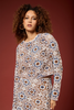 A woman with curly brown hair wearing a long-sleeve knit top and matching skirt featuring an intricate geometric crochet pattern in beige, brown, blue, and white, posed against a solid dark red background.