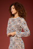 A woman with curly brown hair wearing a long-sleeve dress with an intricate geometric crochet pattern in beige, blue, and brown, posed against a solid dark red background.