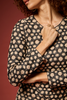 A close-up of a woman wearing a black long-sleeve top with a beige and cream floral pattern, posed against a solid dark red background.
