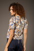 A woman with curly brown hair is shown from the back wearing a short-sleeve cream blouse with an abstract blue and brown pattern, paired with dark blue jeans, against a solid dark gray studio background.
