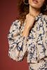 A close-up of a woman wearing a navy and beige floral print blouse with voluminous sleeves and a ruffled front detail, set against a solid dark red background.